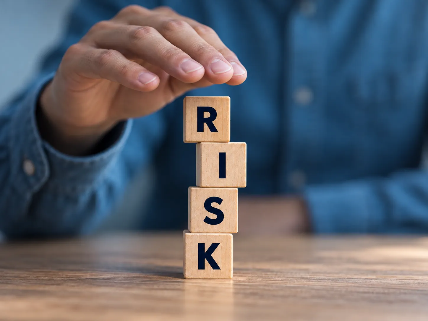 A hand hovering over a stack of four wooden blocks spelling RISK on a wooden table
